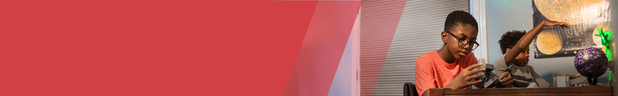 Red section header with two young boys engaged in a hands-on activity at a table. The boy in the foreground wears glasses and a red shirt, focusing intently on assembling a gray block structure. The boy in the background gestures animatedly with his arm raised, with a large space-themed poster and purple model globe visible behind him.