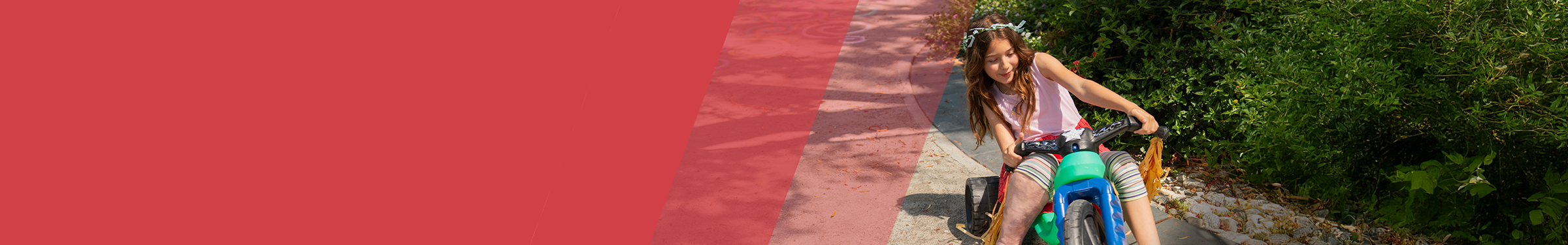 Red section header with young girl to the right with overgrowth on leg riding a colorful tricycle on a sunny path, wearing a flower crown and smiling as she turns the handlebars.